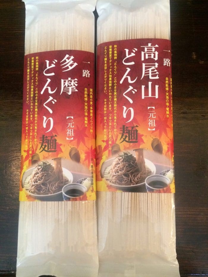 日本の麺類 2 パック。それぞれ赤とオレンジのラベルに、調理済みの麺の入ったボウルと書道の文字が描かれています。パックはダークウッドの表面に並べて置かれ、おいしいスナックと健康食品の両方として、おいしくて栄養のある楽しみを提供します。