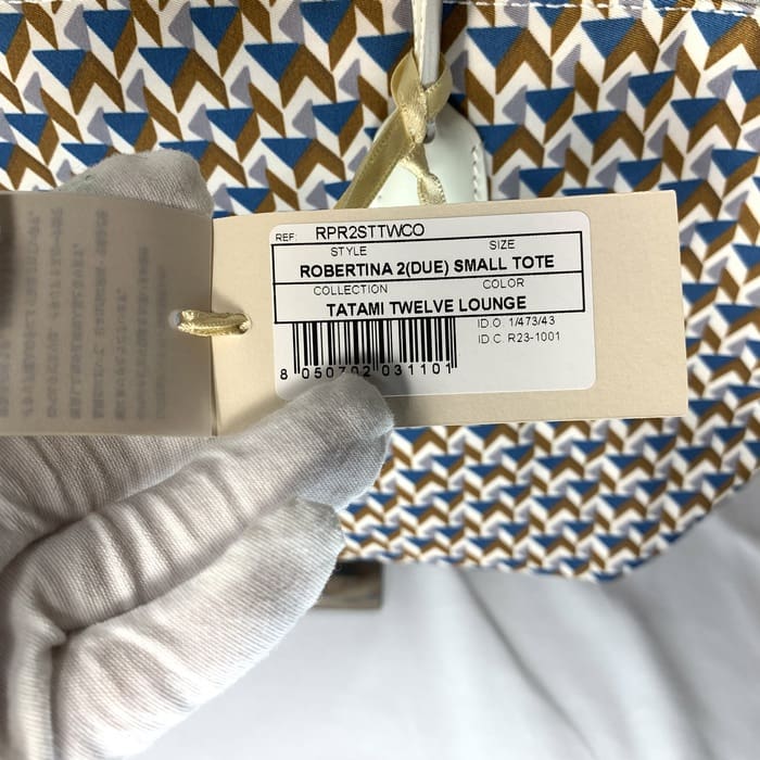 De gehandschoende hand houdt het productlabel "ROBERTINA 2 (DUE) SMALL TOTE" vast op de stof met tatami-patroon met een geometrisch patroon in blauw, bruin en wit, de kenmerkende stijl van de collectie kleine totes van ROBERTA PIERI.