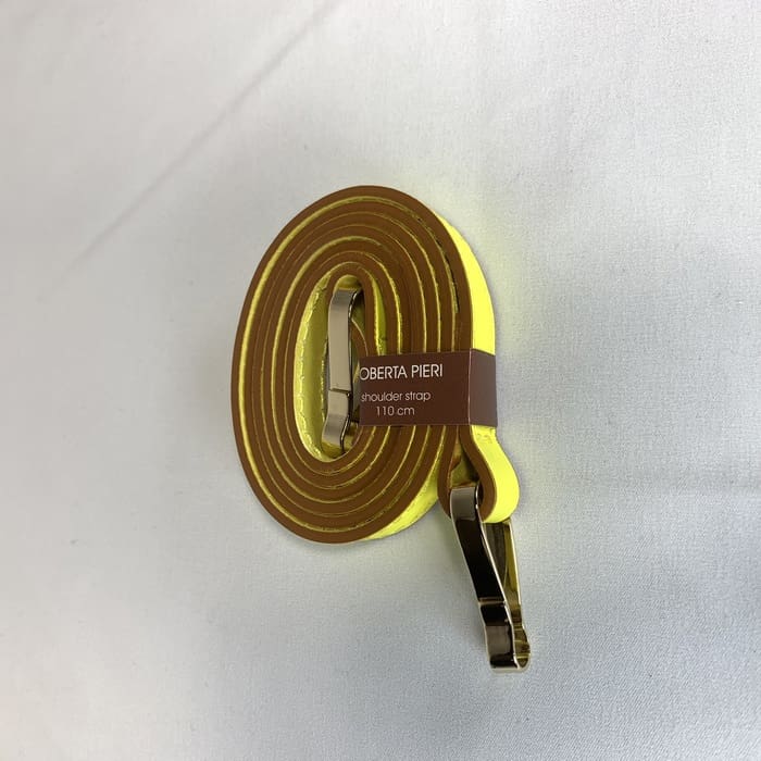 De gele schouderriem met gouden beslag is opgerold en het bruine label "ROBERTA PIERI schouderriem 110 cm" is op het witte oppervlak gesierd.
