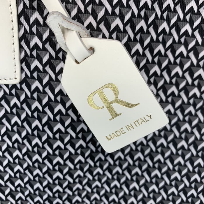 Een wit leren ROBERTA PIERI-label met de letters "R" in goud en de woorden "MADE IN ITALY" hangt aan de achtergrond van een zwart-witte geometrische stof.