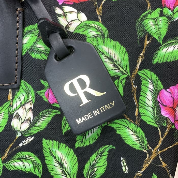 Het zwarte ROBERTA PIERI bagagelabel met de gouden "R" en Italiaanse tekst is opgehangen aan een gebloemde stof met groene bladeren en roze bloemen, als aanvulling op de mini-plunjezak met tijdloze Italiaanse elegantie.