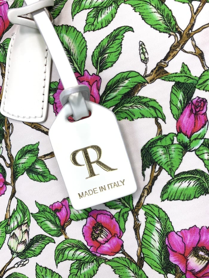 Close-up van ROBERTA PIERI mini plunjezak. steeltjes met groene bladeren en roze bloemen; Het witte label dat aan de 2-way tas is bevestigd, is voorzien van een gouden "R"-logo en de woorden "MADE IN ITALY".