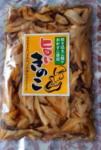 日本語の文字とキノコのイラストが描かれた黄色いラベルの、スライスされたキノコの密封プラスチックパッケージ。「軽井沢のお土産人気商品 旨いキノコ 320g 5袋セット」は、軽井沢で人気の土産品で、贈り物にも、ご家庭で楽しむのにも最適です。.